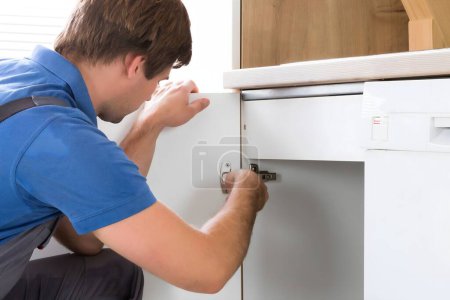 Téléchargez les photos : Réparateur en salopette Réparation Cabinet charnière dans la cuisine - en image libre de droit