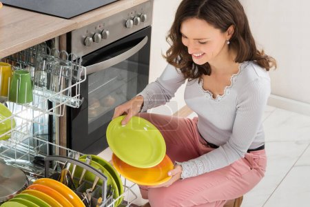 Photo for Young Happy Woman Arranging Plates In Dishwasher At Home - Royalty Free Image