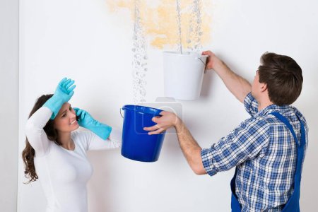 Téléchargez les photos : Jeune femme debout avec travailleur recueillant l'eau dans le seau du plafond dans la maison - en image libre de droit