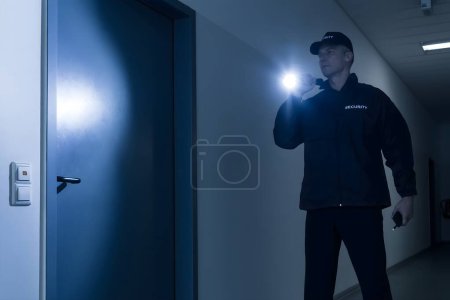 Téléchargez les photos : Gardien de sécurité mature de toute la longueur avec lampe de poche debout devant la porte dans le bâtiment - en image libre de droit