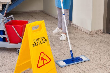 Téléchargez les photos : Jeune ouvrière heureuse avec des équipements de nettoyage et signe de plancher humide sur le plancher - en image libre de droit