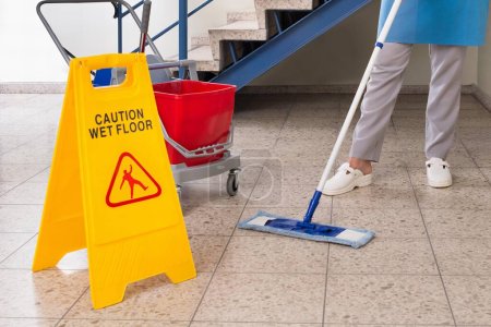 Photo for Young Happy Female Worker With Cleaning Equipments And Wet Floor Sign On Floor - Royalty Free Image