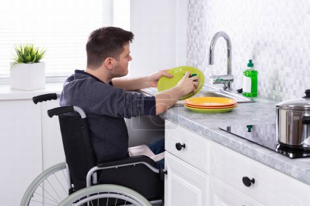 Foto de Hombre sentado en silla de ruedas lavando y limpiando platos en la cocina - Imagen libre de derechos
