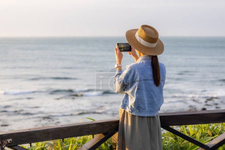 Foto de Viajes mujer utilizar el teléfono celular para tomar fotos en la playa bajo período de puesta del sol - Imagen libre de derechos