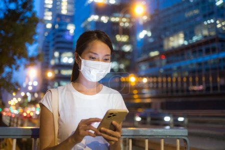 Téléchargez les photos : Femme porter un masque facial et utiliser un téléphone mobile dans la ville la nuit - en image libre de droit