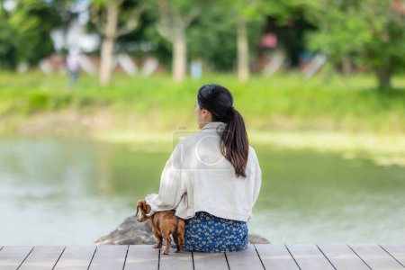 Téléchargez les photos : Femme avec son chien teckel assis sur le sol en bois au bord du lac - en image libre de droit