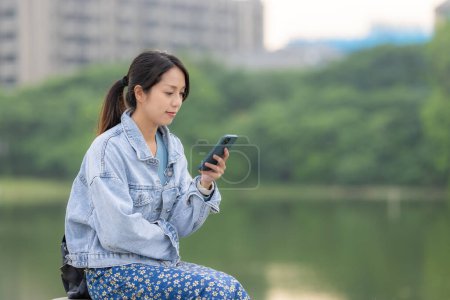 Téléchargez les photos : Femme regardez le téléphone intelligent dans la vue sur le lac au parc du jardin - en image libre de droit