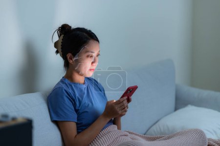 Téléchargez les photos : Femme utiliser un masque en papier et regarder le téléphone portable à la maison le soir - en image libre de droit