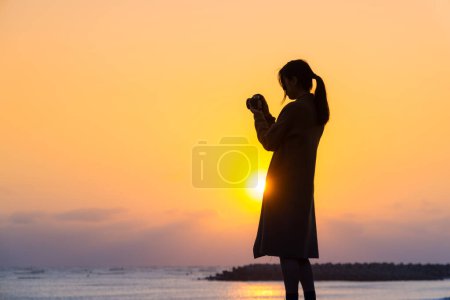 Téléchargez les photos : Silhouette de la femme utiliser un appareil photo pour prendre des photos au coucher du soleil dans la plage - en image libre de droit