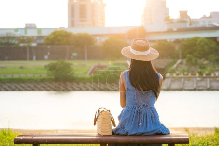 Téléchargez les photos : Femme avec robe et chapeau de paille assis sur le banc au parc sous le coucher du soleil - en image libre de droit
