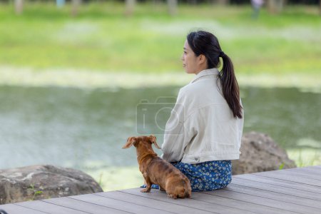 Téléchargez les photos : Femme assis avec son chien teckel au bord du lac dans le parc - en image libre de droit