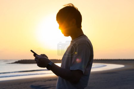 Téléchargez les photos : Silhouette de l'homme utiliser le téléphone mobile au coucher du soleil sur la plage - en image libre de droit
