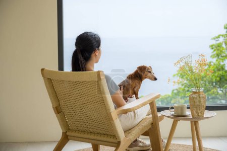Téléchargez les photos : Femme assis sur la chaise avec son chien teckel et profiter de la vue à l'extérieur de la fenêtre - en image libre de droit