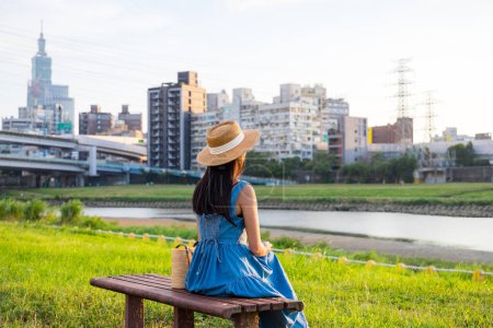 Téléchargez les photos : Femme avec robe et chapeau de paille assis sur le banc au parc sous le coucher du soleil - en image libre de droit