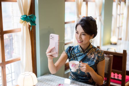 Téléchargez les photos : Femme porter cheongsam chinois traditionnel et l'utilisation du téléphone mobile pour prendre des photos dans le restaurant vintage - en image libre de droit