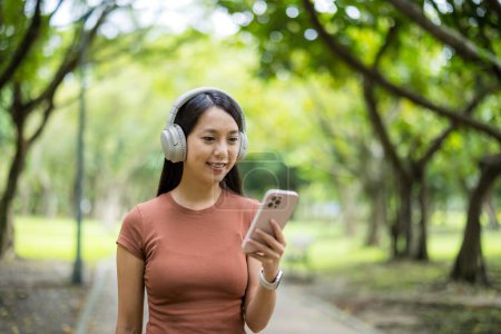 Foto de Mujer usar en los auriculares con el uso de teléfono inteligente caminar a lo largo del parque - Imagen libre de derechos