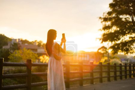 Téléchargez les photos : Femme utiliser le téléphone mobile pour prendre des photos sous le coucher du soleil dans la campagne - en image libre de droit