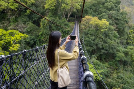 Téléchargez les photos : Femme marcher le long du pont suspendu et utiliser le téléphone portable pour prendre des photos dans la forêt - en image libre de droit