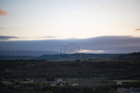 As dusk settles, the soft hues of twilight blanket a serene countryside landscape. The quiet hills and scattered homesteads create a peaceful scene, with the fading light casting a gentle glow over the vast rural expanse