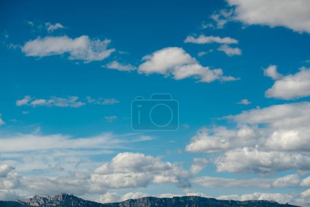 Foto de Nubes blancas esponjosas se dispersan a través de un cielo azul profundo, creando un telón de fondo dramático a los picos escarpados de la montaña visibles en la distancia, haciendo hincapié en la naturaleza majestuosa y expansiva del paisaje. - Imagen libre de derechos