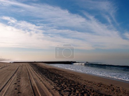 Photo for A low-angle shot of a wide and sandy beach with tire tracks in the sand. The ocean is visible in the background with a rocky jetty and small waves. The sky is blue with wispy clouds. - Royalty Free Image
