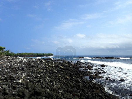 Foto de Una hermosa vista de la costa de Pahoa, Hawaii con cielos azules y olas rompiendo. La foto muestra el contraste entre las rocas negras y la espuma blanca, y la calma del horizonte y el movimiento del agua. La foto está tomada desde un distante - Imagen libre de derechos