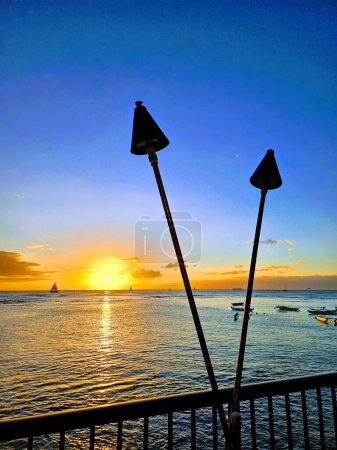 Una vista impresionante de la puesta de sol frente a Waikiki, donde los tonos dorados del sol poniente se fusionan elegantemente con los azules tranquilos del océano. Siluetas antorchas tiki y una barandilla en primer plano añaden un toque de encanto local, enmarcando este sereno mo
