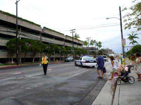 Photo for Honolulu - March 4, 2012: A HPD officer, wearing reflective gear, stands in the middle of a street in Honolulu, Hawaii, to stop traffic due to a water main break. A police car with its lights on is parked diagonally across two lanes of the road. Seve - Royalty Free Image