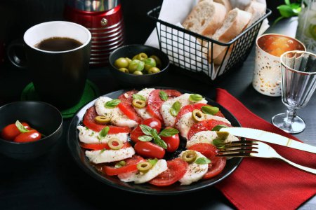 Foto de Ensalada caprese italiana con mozzarella en rodajas y tomates frescos y aceitunas verdes y albahaca en plato negro y mesa - Imagen libre de derechos