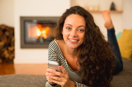 Téléchargez les photos : Belle femme utilisant un téléphone portable à la maison à la chaleur d'une cheminée - en image libre de droit