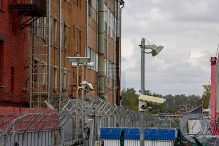 Téléchargez les photos : Barbelé et caméras de surveillance au-dessus de la clôture dans la ville près du mur de construction de briques rouges le jour de l'été, téléobjectif gros plan avec mise au point sélective - en image libre de droit