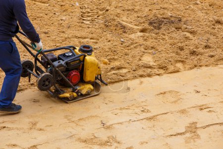 Téléchargez les photos : Travailleur utilisant un compacteur à plaques vibrantes pour le sable de compactage pendant la construction du chemin le jour d'été. - en image libre de droit