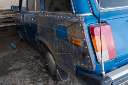 An old blue soviet car with rear left quarter patched with piece of rusted metal mounted with rivets, closeup.