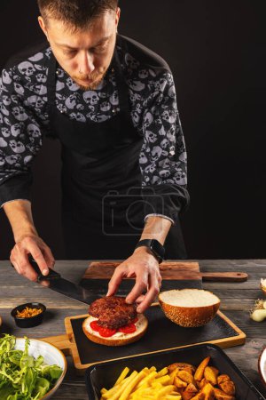 Foto de Chef pone empanada de carne en un pan de hamburguesa. Comida deliciosa y dañina, comida rápida - Imagen libre de derechos