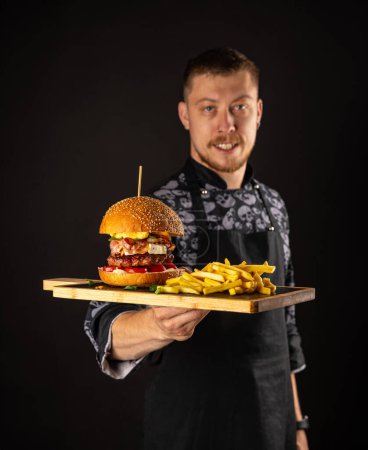 Foto de Chef maestro sonriente en uniforme mirando a la cámara, sosteniendo el plato con hamburguesa servida
. - Imagen libre de derechos
