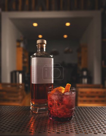Élégant verre de cocktail rouge avec glace et zeste d'orange, à côté de la bouteille de whisky, dans une ambiance de bar confortable