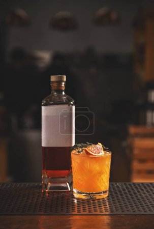 Elegant whiskey cocktail with orange garnish and bottle in a moody bar setting