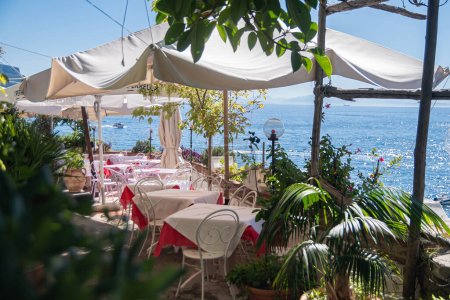 Téléchargez les photos : Café d'été sur la côte de la mer. Tables avec des nappes blanches sous un auvent du soleil chaud de grands buissons verts. Reposez-vous par une chaude journée d'été. Amalfi, Italie, vue sur la mer - en image libre de droit