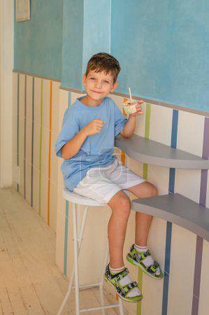 Téléchargez les photos : Mignon garçon de 6 ans dans un T-shirt bleu mange de la crème glacée (gelato) avec une cuillère d'un verre dans une gelateria à Rome. Dessert traditionnel, tourisme avec enfants. Vacances d'été - en image libre de droit