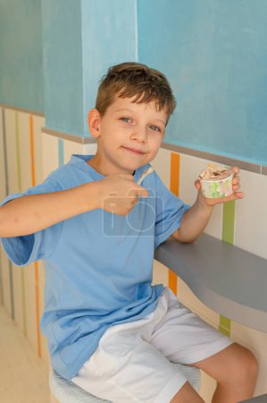 Foto de Niño lindo de 6 años en una camiseta azul come helado (gelato) con una cuchara de un vaso en una gelatería en Roma. Postre tradicional, turismo con niños. Vacaciones de verano - Imagen libre de derechos