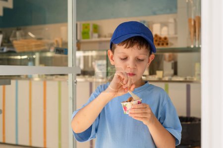 Foto de Niño lindo de 6 años en una camiseta azul come helado (gelato) con una cuchara de un vaso en una gelatería en Roma. Postre tradicional, turismo con niños. Vacaciones de verano - Imagen libre de derechos