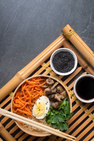 Photo for Noodle soup, ramen in a white bowl (bowl) with chicken egg, carrots, mushrooms on a bamboo tray on a dark stone background. Asian traditional fast food - Royalty Free Image