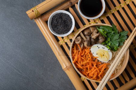 Téléchargez les photos : Soupe de nouilles, ramen dans un bol blanc (bol) avec oeuf de poulet, carottes, champignons sur un plateau de bambou sur un fond de pierre sombre. Asiatique traditionnel fast food - en image libre de droit