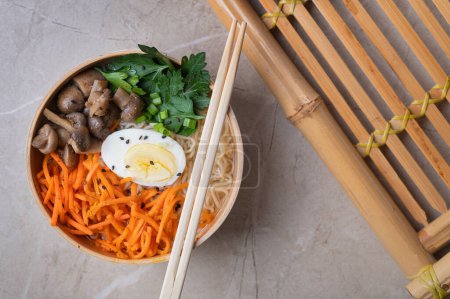 Téléchargez les photos : Soupe de nouilles, ramen dans un bol blanc (bol) avec oeuf de poulet, carottes, champignons sur un plateau de bambou sur un fond de pierre claire. Asiatique traditionnel fast food - en image libre de droit