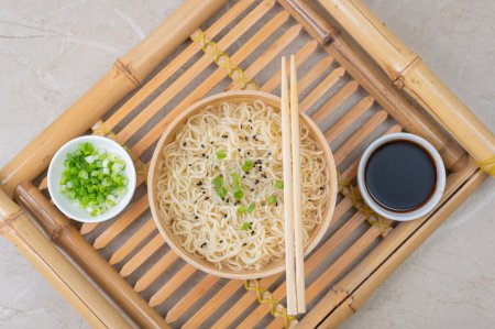 Téléchargez les photos : Assiette en bambou blanc (bol) avec nouilles aux œufs et assiettes avec ingrédients pour elle sur un plateau de service en bambou sur un fond clair. Asiatique traditionnel fast food - en image libre de droit