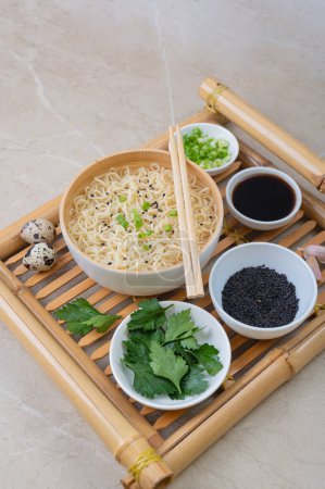 Téléchargez les photos : Assiette en bambou blanc (bol) avec nouilles aux œufs et assiettes avec ingrédients pour elle sur un plateau de service en bambou sur un fond clair. Asiatique traditionnel fast food - en image libre de droit