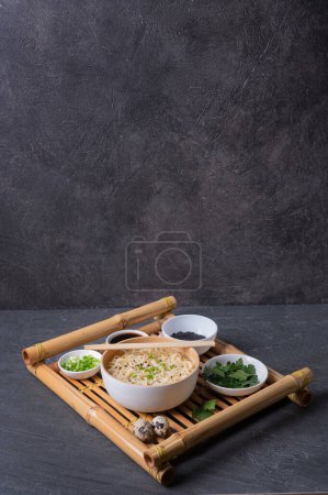 Téléchargez les photos : Assiette en bambou blanc (bol) avec nouilles aux œufs et assiettes avec ingrédients pour elle sur un plateau de service en bambou sur un fond en marbre foncé (ardoise). Asiatique traditionnel fast food - en image libre de droit