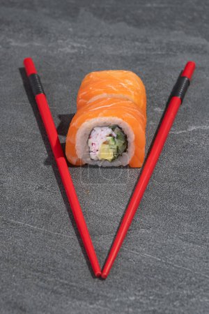 Téléchargez les photos : Rouleaux de sushi au saumon, avocat et concombre avec baguettes rouges sur fond de pierre noire (ardoise). Alimentation traditionnelle asiatique. - en image libre de droit