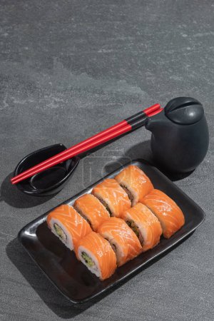 Téléchargez les photos : Les sushis roulent avec du saumon, de l'avocat et du concombre sur une assiette rectangulaire noire sur un fond de pierre noire (ardoise). Alimentation traditionnelle asiatique. - en image libre de droit