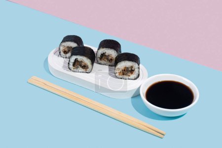 Téléchargez les photos : Hosomaki (sushi) avec anguille sur un stand blanc sur un fond uni coloré (bleu, rose). Une composition simple et concise - en image libre de droit